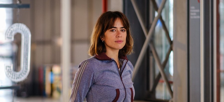 Émilie Tronche, dessinatrice connue notamment pour sa série « Samuel », pose avec assurance dans un espace emblématique du Centre Pompidou. Vêtue d’un haut de survêtement violet chiné aux détails bordeaux et d’un jean taille haute, elle s’appuie légèrement contre une rambarde blanche en métal ajouré. Son regard franc et tranquille est dirigé vers l’objectif, tandis que la lumière ambiante met en valeur ses traits et son style sobre et contemporain. En arrière-plan, les structures métalliques croisées et les parois vitrées caractéristiques du bâtiment dialoguent avec une grande lettre « C » suspendue et lumineuse, renforçant l’ambiance artistique et architecturale du lieu. Cette photo, prise par Pierre Malherbet, capte un instant d’intimité dans un cadre emblématique de la scène culturelle parisienne.