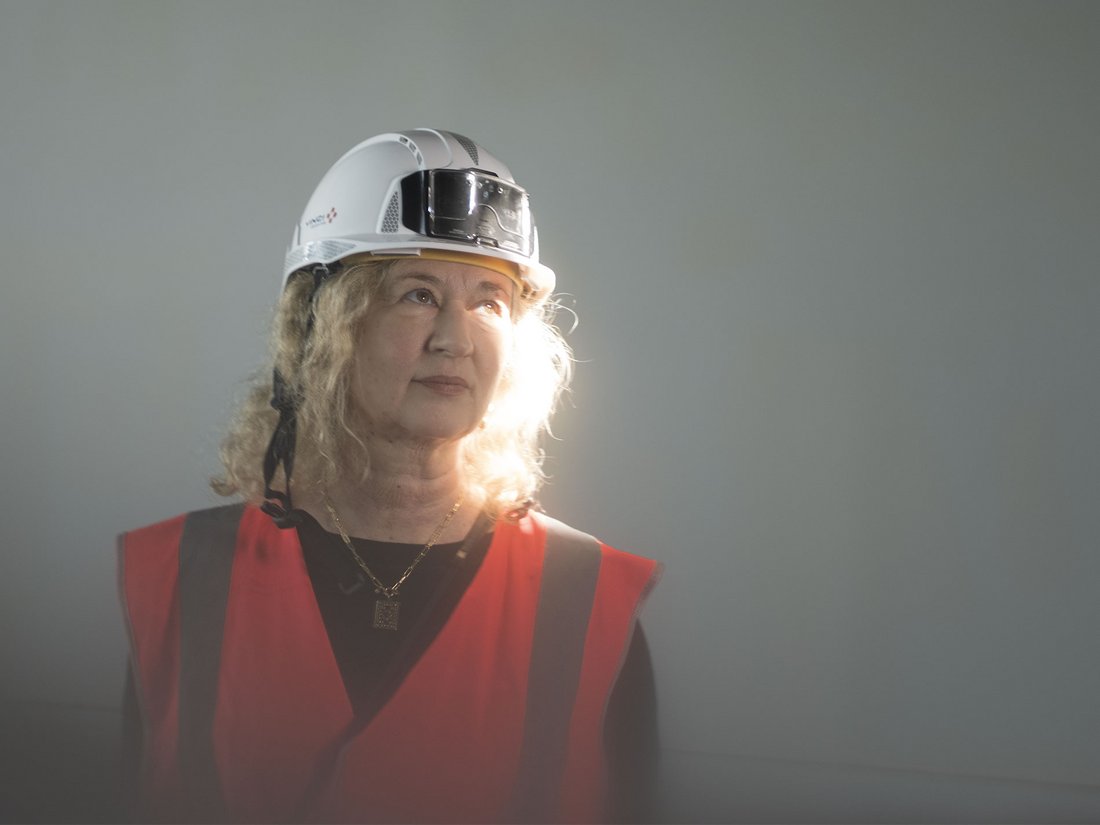 Portrait d’Alexia Fabre au Centre Pompidou Francilien encore en travaux. Elle porte un casque de chantier blanc et un gilet de sécurité orange. Une vive lumière éclaire son visage tandis qu’elle regarde vers le haut.