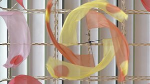 Dans l’immense nef lumineuse du Grand Palais à Paris, l’installation « Voûtes et volutes » flotte avec légèreté sous le plafond métallique vert pâle. Suspendue à une structure géométrique en bois clair, un ensemble de fanions en soie ultra fine se déploie dans l’espace, évoquant des courbes aériennes, des flux d’air ou des mouvements chorégraphiques. Les textiles, aux couleurs vibrantes – rouge, violet, jaune, rose orangé – sont ponctués de cercles colorés, jouant avec la transparence de la matière. Grâce à des moteurs dissimulés dans la structure, les éléments s’animent doucement, créant des ondulations et des spirales hypnotiques. L’ensemble donne vie à un ballet suspendu, en perpétuelle transformation, où la légèreté de la soie contraste avec la rigueur de l’ossature en bois. L’espace épuré de la galerie amplifie la sensation de flottement et d’apaisement, invitant les visiteurs à lever les yeux et à se laisser porter par ce paysage en suspension.