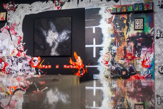 This large-scale mural dominates an entire wall, blending street art, abstraction, and visual storytelling. On the left, a collage of black-and-white newspaper clippings forms the background, layered with expressive, screen-printed faces and splashes of red and orange paint. In the center, a framed black and grey abstract painting evokes a stormy or geological form, full of shadows and texture. To the right, a chaotic painted area bursts with vivid colors, fragmented textures, scorched effects, graffiti, and overlapping images—like fragments of urban memory. A small grid of photos or prints evokes a sense of catalogued memories. Fluorescent splashes and torn visuals energize the composition. The soft lighting and reflective floor add depth, mirroring parts of the mural and enhancing its immersive quality.