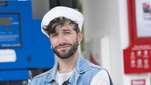 Guillaume Blot, photographe français au style affirmé, pose devant une station-service. Gilet en jean, béret blanc et banane en bandoulière composent un look urbain et décontracté.