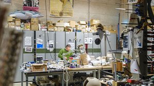 L’image montre un vaste atelier foisonnant d’énergie créative, où un artisan ou technicien en pull vert se penche sur un plan de travail encombré d’outils, de câbles et de composants électroniques. Les étagères et les armoires métalliques à l’arrière débordent de cartons et de matériaux, témoignant d’une activité intense et expérimentale. Au premier plan, des roues de vélo, des pinces et des boîtes d’outils soulignent la dimension manuelle et technique du lieu. Les affiches artistiques accrochées au mur ajoutent une touche culturelle, révélant un espace hybride entre art, invention et artisanat industriel.