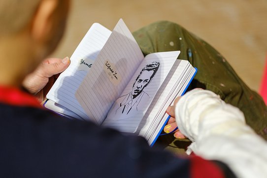 Shuck One, a seminal figure in street art, is shown here in a photograph by Pierre Malherbet, intently examining a sketchbook open across his lap. Captured from behind, the image highlights his hands delicately holding a lined notebook that reveals a black line drawing: a portrait of a man with an intense gaze and elegantly styled hair. On the facing page, handwritten notes can be discerned, including the name “Wifredo Lam” alongside various dates.  The neutral, softly blurred background draws the viewer’s focus to the sketchbook and the artist’s gestures. Dressed in a black-and-red striped sweater and paint-stained trousers—a testament to his immersion in the creative process—Shuck One appears absorbed in a moment of reflection or research. This intimate scene unveils a contemplative phase in his practice, intertwining memory, preparatory drawing, and graphic homage.