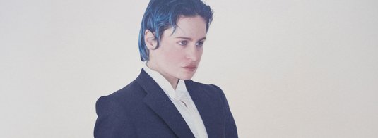 Portrait en plan moyen de Rahim C. Redcar, alias Christine and the Queens, vêtu d’un costume noir structuré sur une chemise blanche immaculée. Son regard intense, dirigé hors champ, et sa posture soignée traduisent une présence à la fois théâtrale et introspective. Ses cheveux bleu foncé, plaqués vers l’arrière, apportent une touche moderne et audacieuse à cette silhouette androgyne. Le fond uni, clair, renforce la sobriété de la composition et met en lumière l’esthétique singulière de l’artiste, entre élégance classique et affirmation identitaire.