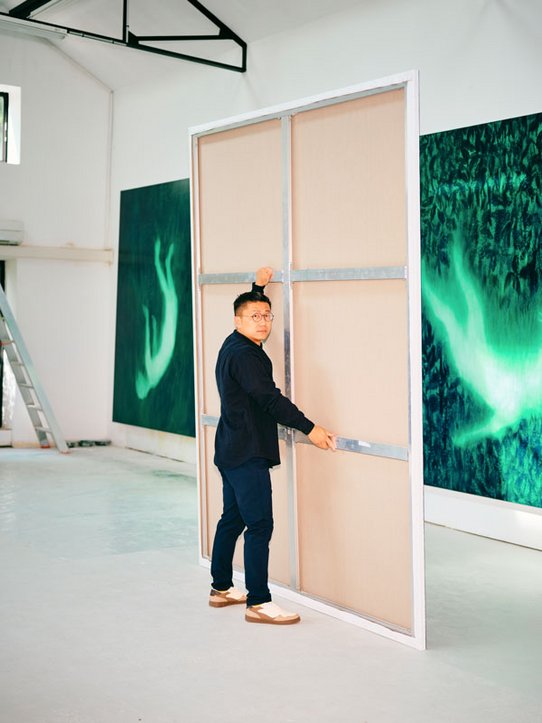 L’artiste Xie Lei, vêtu d’une veste sombre et de chaussures claires, déplace une grande toile dans son atelier lumineux. En arrière-plan, d’immenses peintures aux teintes vertes et sombres représentent des formes évanescentes, presque spectrales, donnant une atmosphère à la fois mystérieuse et poétique.