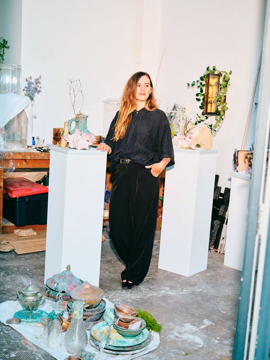 Bianca Bondi, debout dans son atelier, vêtue de noir, pose entre deux socles blancs exposant des compositions organiques et sculpturales. Autour d’elle, des objets patinés, des vases et des installations mêlant nature et art créent une atmosphère à la fois mystique et contemporaine.