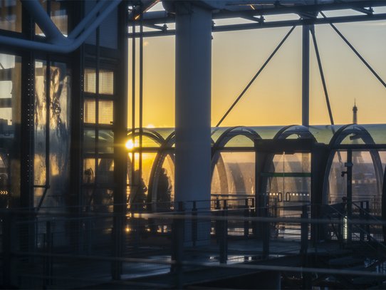 Cette photographie de Pierre Malherbet révèle une vision graphique et lumineuse du Centre Pompidou à Paris au coucher du soleil. Les structures métalliques emblématiques, les arches vitrées et les passerelles industrielles se découpent dans une lumière dorée qui traverse l’architecture contemporaine avec douceur. Les reflets sur les surfaces transparentes créent un jeu subtil entre ombre et lumière, renforçant la profondeur et l’élégance de la composition. À l’arrière-plan, la silhouette discrète de la tour Eiffel apparaît dans la brume lumineuse, ajoutant une dimension typiquement parisienne à la scène. L’atmosphère est calme, presque cinématographique, mêlant modernité urbaine, design architectural et poésie visuelle. Cette image met en valeur l’esthétique industrielle du Centre Pompidou tout en capturant la chaleur et la sérénité de la lumière de fin de journée.