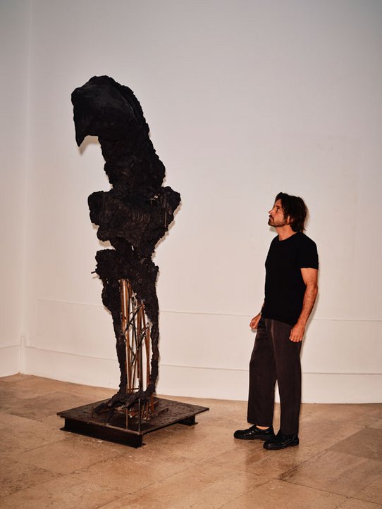 Lionel Sabatté lors du montage de son exposition. La première photo le montre face à une sculpture monumentale sombre, aux formes organiques, presque anthropomorphiques. La seconde le capture en train de travailler une sculpture miniature en poussière et matériaux bruts, posée sur un socle blanc. Deux échelles d’échelle – l’intime et le monumental – se répondent dans sa démarche.