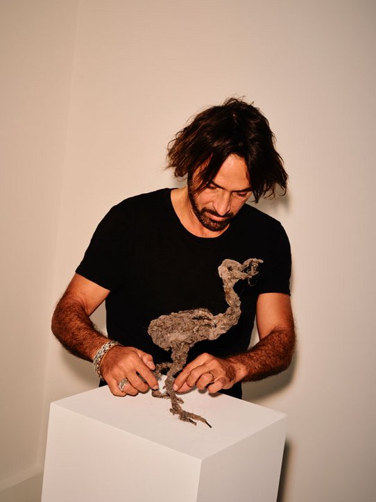 Lionel Sabatté lors du montage de son exposition. La première photo le montre face à une sculpture monumentale sombre, aux formes organiques, presque anthropomorphiques. La seconde le capture en train de travailler une sculpture miniature en poussière et matériaux bruts, posée sur un socle blanc. Deux échelles d’échelle – l’intime et le monumental – se répondent dans sa démarche.