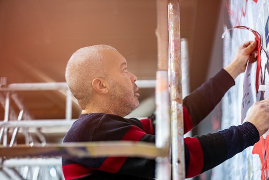 Urban artist Shuck One, photographed by Pierre Malherbet, is captured mid-action, fully absorbed in the creation of a mural. Standing on scaffolding, arms raised, he carefully applies a graphic element to the wall. His gaze is steady and focused, revealing his total commitment to the artistic gesture. Dressed in a sweater with broad black and burgundy stripes, he stands out slightly from the blurred yet colorful background, where splashes of paint and abstract forms can be discerned. Beside him, the metal structure of the scaffolding evokes the technical and physical demands of mural work. The ambient light—soft and tinged with orange—enhances the atmosphere of intense, silent creation, as if suspended in time. This scene reflects the artist’s physical and emotional engagement in his creative process.