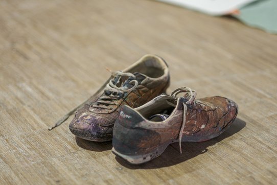 Casually set on a wooden floor, this worn pair of shoes bears the visible marks of time and creation. Spattered with multicoloured paint—shades of violet, red, ochre, and black—the battered sneakers speak to the daily practice of the artist Shuck One. Each stain and smudge appears to narrate a fragment of his artistic process, like a silent extension of his gestures on canvas or wall.  The worn soles, untied laces, and weathered leather texture reinforce a sense of raw authenticity and lived experience. This minimalist yet evocative image encapsulates the materiality of urban art: work that unfolds on the ground, at heights, in motion—work into which the artist fully immerses himself. More than a mere accessory, these shoes become a discreet yet powerful imprint of Shuck One’s creative path.
