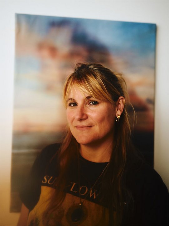 Portrait chaleureux de l'artiste Eva Nielsen, souriante, debout devant une œuvre aux teintes pastel représentant un ciel flou et coloré.