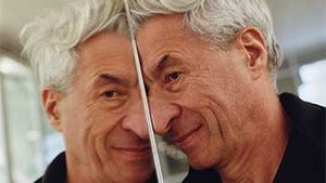 Ce portrait présente l’artiste contemporain Maurizio Cattelan, penché doucement contre une surface miroir qui coupe verticalement l’image en deux. Son visage, capturé en double reflet, affiche un demi-sourire ironique, signature de son univers provocateur et empreint d’humour. Ses cheveux gris décoiffés encadrent un regard vif et joueur. Vêtu d’un blouson beige clair avec une doublure noire, il pose dans un environnement lumineux et légèrement flouté, probablement une galerie ou un espace d’exposition. Le miroir, en divisant le visage de Cattelan, évoque ses thèmes récurrents de la duplicité, de l’identité et du regard critique sur l’art lui-même.