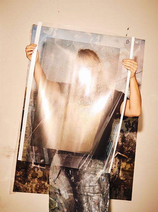 ’artiste Eva Nielsen tient devant elle une œuvre semi-transparente, masquant partiellement son visage, dans un atelier aux murs neutres tapissés de paysages texturés et picturaux.
