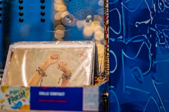 Cette photographie présente un détail percutant de l’installation de Valérie John. En avant-plan, une boîte cartonnée, réutilisée et décorée, contient une image protégée sous plastique représentant deux bras levés, poings serrés, encore partiellement enchaînés. Ce geste puissant, universellement reconnu comme un symbole de résistance, de libération ou de lutte anticoloniale, est amplifié par le contexte visuel et symbolique de l’œuvre. Le fond est occupé par un mur bleu indigo orné de motifs graphiques évoquant des écritures, des signes ou des vagues, sur lequel se reflètent des guirlandes lumineuses dorées. Ce contraste entre lumière festive et image de combat donne à la scène une tension particulière, où la mémoire de l’esclavage et des luttes pour la dignité humaine s’entrelace à l’esthétique. Ce fragment de l’installation SECRET(S)... RÊVES DE PAYS... FABRIQUE À MÉMOIRE(S)... PALIMPSESTE... (1998–2025), met en lumière le rôle central du corps et des gestes dans les récits postcoloniaux, tout en inscrivant l’objet photographique dans une démarche de recyclage, de détournement et de sacralisation populaire.