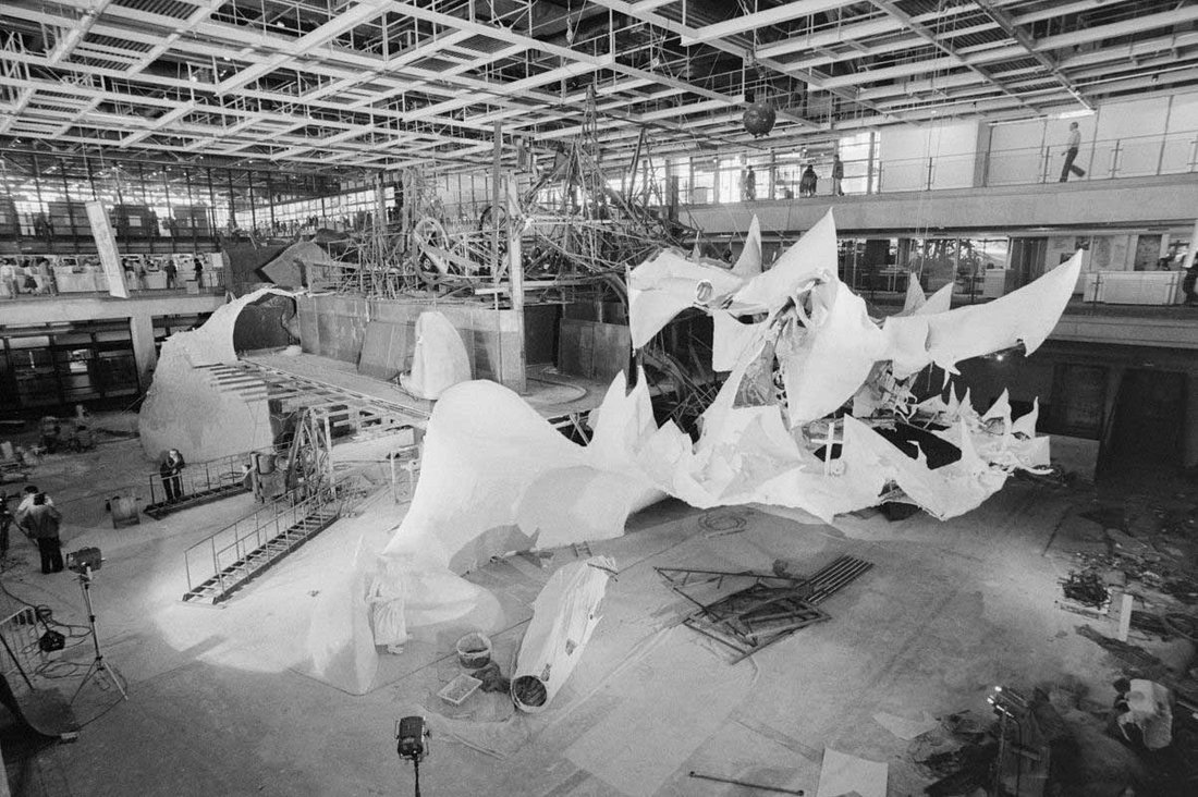 Cette photographie en noir et blanc documente une étape clé de la construction monumentale du « Crocrodrome de Zig et Puce » par Jean Tinguely, dans le Forum du Centre Georges Pompidou, en 1977. L’image révèle l’ampleur colossale de l’installation, occupant tout le volume central du musée avec une structure mêlant acier, bois, tissu, résine et éléments cinétiques. À gauche, une plateforme surélevée en cours de montage est reliée à un ensemble sculptural composé de tunnels et de formes arrondies évoquant des carcasses organiques ou animales. Au centre, une structure métallique dense — constituée de poutres, de roues dentées, de câbles et de mécanismes articulés — illustre la signature visuelle de Tinguely, entre chaos mécanique et poésie de la machine. À droite, d’immenses pans de matière souple et déchiquetée flottent ou pendent, évoquant la mâchoire ouverte d’un monstre, avec ses crocs déployés dans l’espace. Au sol, outils, projecteurs, échafaudages et matériaux jonchent la zone de travail, témoignant de l’aspect performatif du montage. Quelques silhouettes humaines donnent l’échelle : minuscules face à cette œuvre immersive. En arrière-plan, les visiteurs curieux observent depuis les passerelles vitrées du musée, soulignant l’interaction entre art, architecture et public.