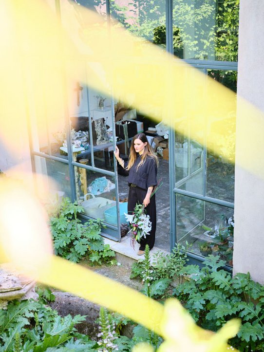 Bianca Bondi se tient sur le seuil de son atelier baigné de lumière, un bouquet de fleurs en main, entourée d'une végétation luxuriante. À travers les grandes vitres, on devine l'intérieur de son espace de création, empli d’objets artistiques et de matériaux en cours d’usage.