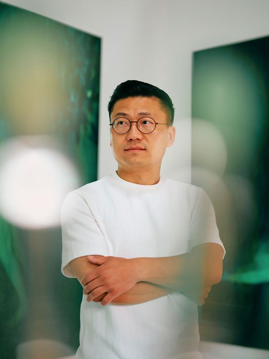 Xie Lei, artiste contemporain, pose les bras croisés dans son atelier lumineux. Vêtu d’un t-shirt blanc et de lunettes rondes, il se tient devant ses grandes toiles aux teintes vertes et sombres, qui diffusent une atmosphère mystérieuse et onirique.