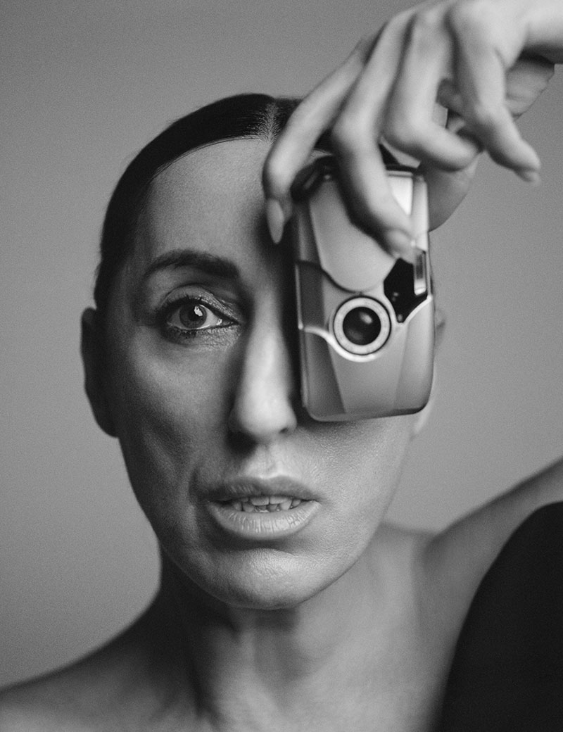 Portrait noir et blanc au style éditorial et minimaliste. Une femme au visage fortement cadré regarde l’objectif avec intensité, tandis que sa main levée place un petit appareil argenté devant un œil, créant un jeu visuel entre corps, technologie et identité. La lumière douce souligne la texture de la peau, les lèvres entrouvertes et le maquillage lumineux. Le fond neutre renforce l’esthétique contemporaine, presque sculpturale, entre photographie de mode, art conceptuel et accessoire futuriste.