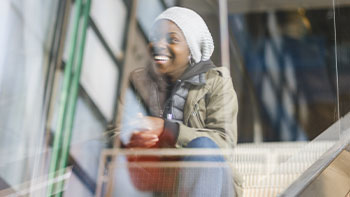 Josza Anjembe, réalisatrice franco-camerounaise, monte un escalator vitré du Centre Pompidou. En arrière-plan, les structures tubulaires emblématiques du bâtiment se dessinent sous une lumière douce. Son expression calme et assurée reflète la créativité et la détermination. L’ambiance urbaine et artistique du lieu amplifie la portée culturelle de la scène.