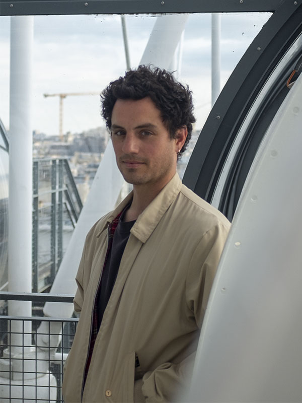 Aristide Barraud, ancien rugbyman devenu poète et auteur, se tient dans une passerelle vitrée du Centre Pompidou. Vêtu d’un long manteau beige, il regarde l’objectif avec calme. Autour de lui, les tubes métalliques emblématiques du bâtiment contrastent avec le ciel nuageux. En arrière-plan, une grue et les toits parisiens suggèrent un Paris en mutation. La scène mêle poésie urbaine et architecture moderne.