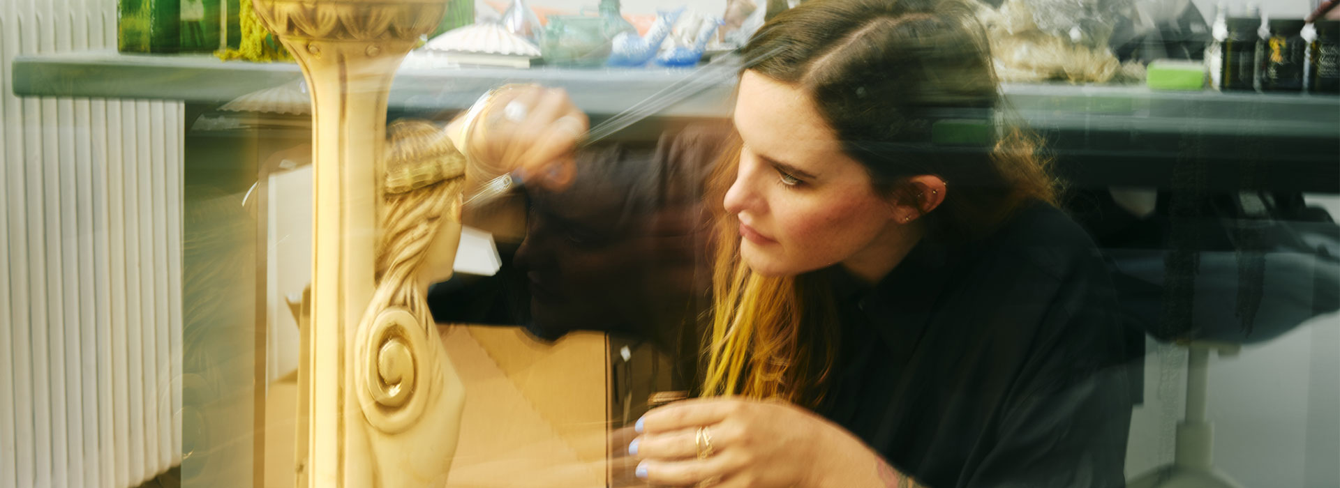 Une artiste, Bianca Bondi, agenouillée dans un atelier, sculpte ou restaure une colonne décorative ornée d’un serpent doré, entourée d’objets artistiques, de fleurs séchées et d’éléments naturels.