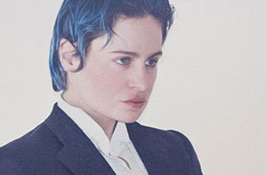 Mid-shot portrait of Rahim C. Redcar, also known as Christine and the Queens, wearing a structured black suit over a crisp white shirt. His intense, off-camera gaze and composed posture convey both theatrical presence and deep introspection. The slicked-back dark blue hair adds a bold, modern edge to his androgynous silhouette. The plain, light background enhances the minimalist elegance and draws focus to the unique identity and artistic style of the performer, blending classic formality with personal expression.