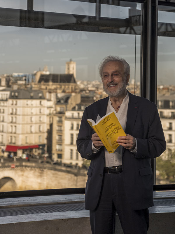 Claude Mollard, vêtu d’un élégant costume sombre et d’une chemise blanche ouverte, pose souriant dans une lumière dorée de fin de journée. Il tient un livre jaune vif intitulé "L'Épopée Beaubourg". À travers la baie vitrée, on distingue les toits parisiens baignés de lumière et un pont de pierre enjambant la Seine. L’ambiance est à la fois intellectuelle, détendue et contemplative, capturant un instant de réflexion culturelle en plein cœur de Paris.