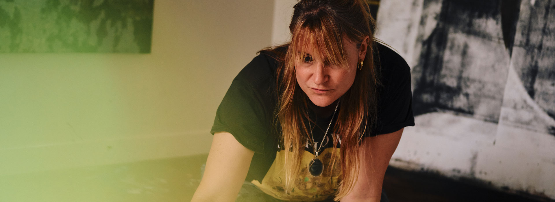 Eva Nielsen, artiste contemporaine, agenouillée dans son atelier, concentrée sur une œuvre au sol, entourée de peintures abstraites, avec un filtre lumineux flou au premier plan.