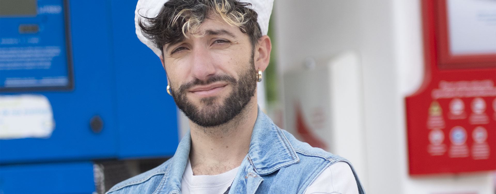 Guillaume Blot, photographe français au style affirmé, pose devant une station-service. Gilet en jean, béret blanc et banane en bandoulière composent un look urbain et décontracté.