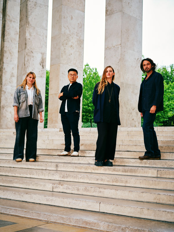 Sur les marches de pierre du musée d’Art moderne de Paris, quatre artistes finalistes du prix Marcel Duchamp posent avec assurance. À gauche, Eva Nielsen en tenue décontractée, puis Xie Lei bras croisés, Bianca Bondi au regard calme et Lionel Sabatté en veste sombre. L’arrière-plan de colonnes massives et de feuillages verts confère à la scène une dimension solennelle et artistique.