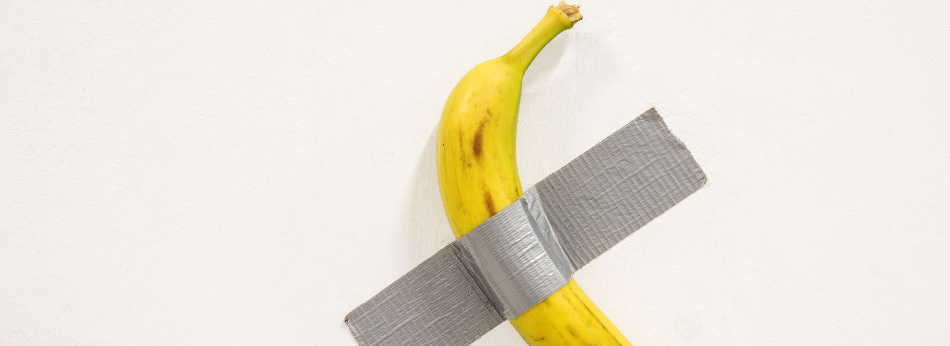 Une banane jaune tachetée de brun, bien mûre, est scotchée de manière brute contre un mur blanc uni. Elle est maintenue par un large morceau de ruban adhésif gris disposé en biais, créant une croix imparfaite. L'œuvre joue volontairement sur la simplicité et l’absurde, évoquant une esthétique minimaliste et provocante. L’absence d’arrière-plan ou d’élément décoratif met en valeur la nature décalée de l’objet, emblématique de l’art contemporain conceptuel. Cette image fait directement référence à l’œuvre « Comedian » de Maurizio Cattelan, célèbre pour sa critique de la marchandisation de l’art et sa capacité à susciter débat et réflexion à partir d’un objet banal.