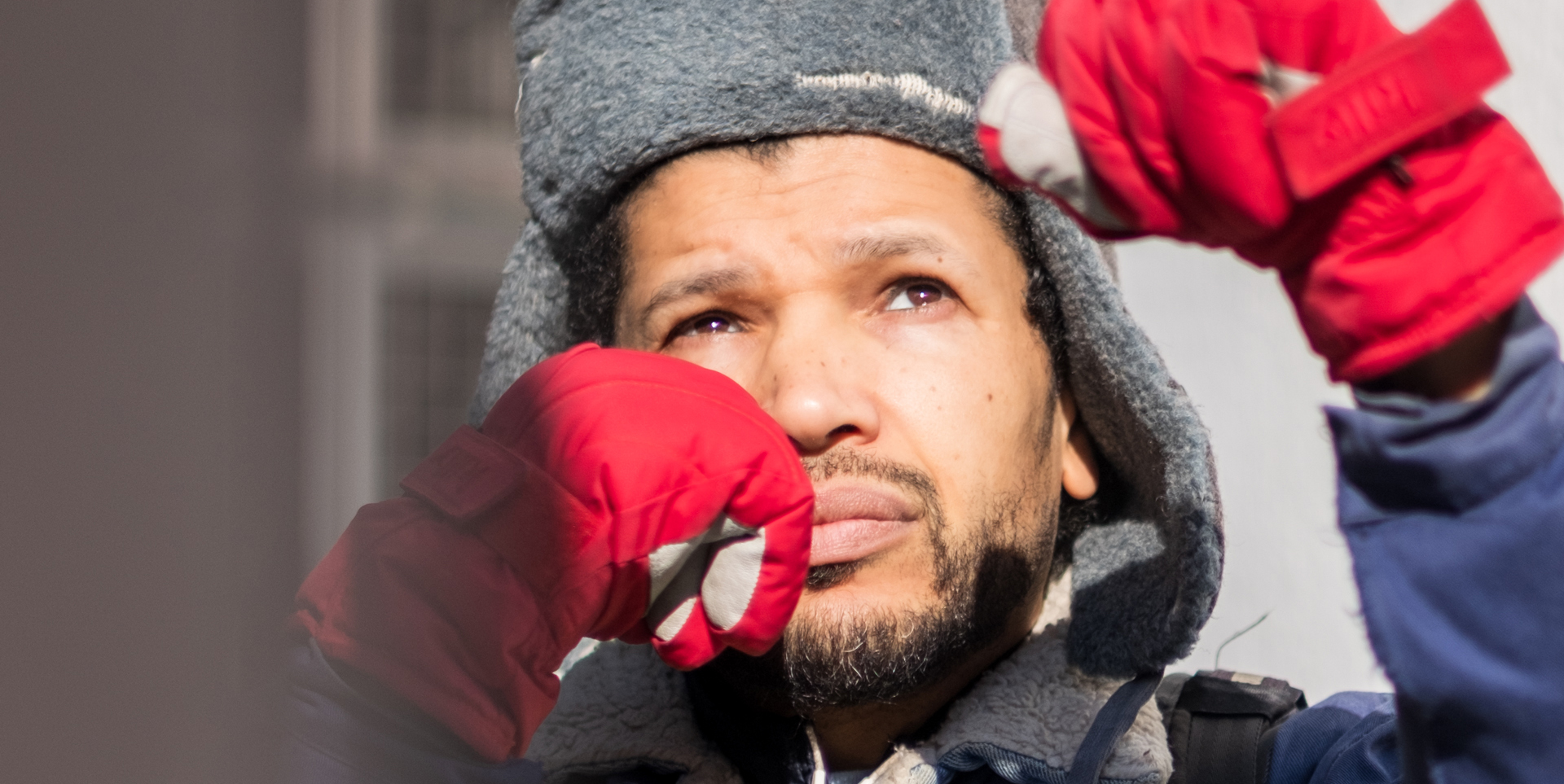 Un homme en gros plan est photographié dans une lumière crue et hivernale, le visage tourné vers le haut avec une expression à la fois concentrée et contemplative. Il porte un bonnet épais en laine grise à oreillettes, typique des climats froids, et des gants d’un rouge vif qui attirent immédiatement l’attention. Ses mains sont levées à hauteur du visage, suggérant une action précise ou une gestuelle symbolique. Sa veste bleu foncé à doublure en laine claire évoque un style utilitaire, robuste. L’arrière-plan flou laisse deviner une structure urbaine ou industrielle, renforçant l’ambiance de travail ou d’engagement dans un environnement extérieur. Le contraste entre la lumière vive et les ombres profondes souligne la texture de sa peau, la densité de la laine, et l’intensité de son regard.