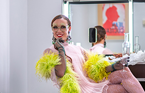Dans une loge foisonnante de couleurs, d’accessoires et d’énergie créative, Romain Brau photographié par Pierre Malherbet se dévoile dans toute sa flamboyance. Installé avec décontraction sur un fauteuil recouvert de fausse fourrure beige, il sourit avec connivence tout en tenant un verre d'eau. Son look exubérant mêle une robe fluide rose poudré à des collants en dentelle rose, rehaussé d’un somptueux boa en plumes jaune citron et vert émeraude. De larges lunettes rondes, un maquillage pailleté prononcé, des lèvres carmin et des bijoux imposants complètent son allure de diva glamour. Autour de lui, des vêtements éparpillés et des accessoires détonants accentuent l’atmosphère de coulisses animées. Dans le miroir rétro-éclairé derrière lui, son reflet ajoute une dimension performative à la scène. Et surtout, au mur, un portrait de Jean Cocteau — identifiable à son style caractéristique — vient ancrer cette scène de vie contemporaine dans une lignée artistique iconique, entre modernité queer et héritage culturel.