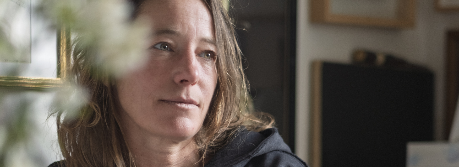 Ce portrait de Séverine Hubard, artiste plasticienne reconnue pour ses installations monumentales et ses œuvres in situ, est capturé dans un cadre intime. Assise en intérieur, elle est vêtue d’un sweat noir à capuche et entourée de cadres accrochés au mur, d’objets personnels et de flous végétaux au premier plan. Son regard, tourné vers la gauche de l’image, dégage une expression à la fois douce et méditative, empreinte de calme. La lumière naturelle venant de côté éclaire subtilement son visage, mettant en valeur la texture de sa peau et la légèreté de ses cheveux détachés. Cette photographie, bien que simple, transmet une forte présence humaine et artistique, révélant un moment de pause ou de réflexion dans l’univers créatif de l’artiste.