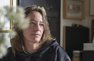 Ce portrait de Séverine Hubard, artiste plasticienne reconnue pour ses installations monumentales et ses œuvres in situ, est capturé dans un cadre intime. Assise en intérieur, elle est vêtue d’un sweat noir à capuche et entourée de cadres accrochés au mur, d’objets personnels et de flous végétaux au premier plan. Son regard, tourné vers la gauche de l’image, dégage une expression à la fois douce et méditative, empreinte de calme. La lumière naturelle venant de côté éclaire subtilement son visage, mettant en valeur la texture de sa peau et la légèreté de ses cheveux détachés. Cette photographie, bien que simple, transmet une forte présence humaine et artistique, révélant un moment de pause ou de réflexion dans l’univers créatif de l’artiste.