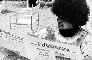 Assise à une terrasse extérieure jonchée de graviers, Sarah Maldoror lit avec attention un exemplaire du journal "l'Humanité". Elle porte une robe à motifs floraux accompagnée d’un col en dentelle blanche délicate, et un sous-pull noir. Sa coiffure afro dense encadre son visage concentré, dirigé vers l’article principal dont le titre en Une est : « Les artifices ont fait long feu ». L’environnement est calme, avec des chaises et tables en métal blanc typiques des cafés ou lieux publics en plein air, légèrement vides, ce qui accentue une impression de sérénité ou de moment volé dans la routine. Le cliché en noir et blanc ajoute une dimension documentaire et intemporelle, évoquant une époque de militantisme ou de réflexion intellectuelle engagée, en lien avec la nature politique du journal lu.
