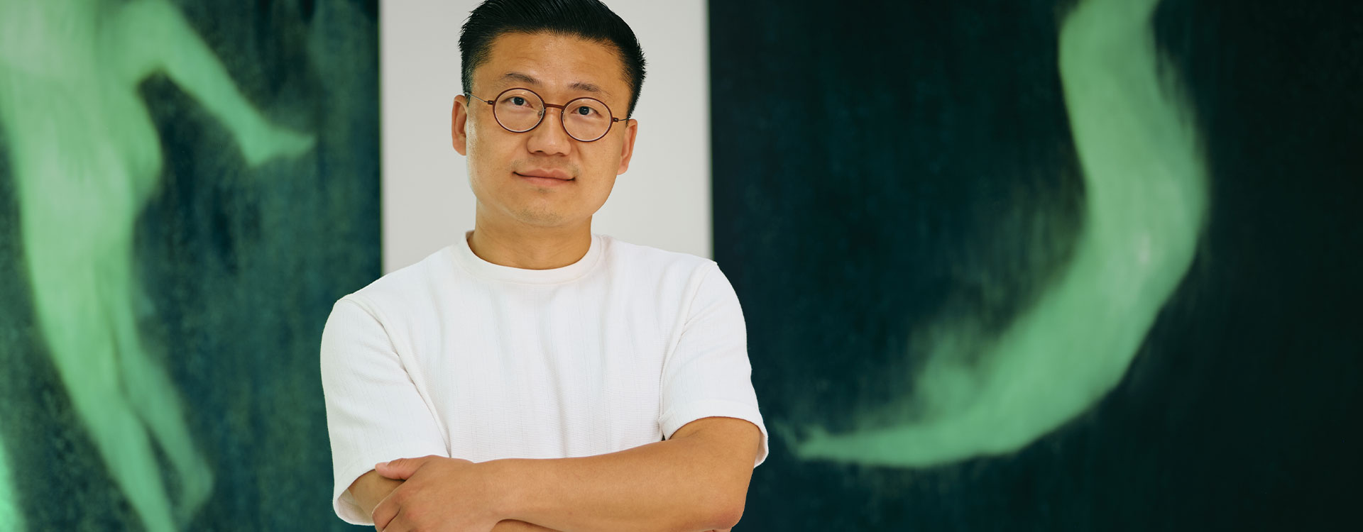 ie Lei, artiste contemporain, pose dans son atelier lumineux. En arrière-plan, deux grands tableaux sombres révèlent des silhouettes vertes spectrales, semblant flotter ou tomber, créant une ambiance onirique et mystérieuse.