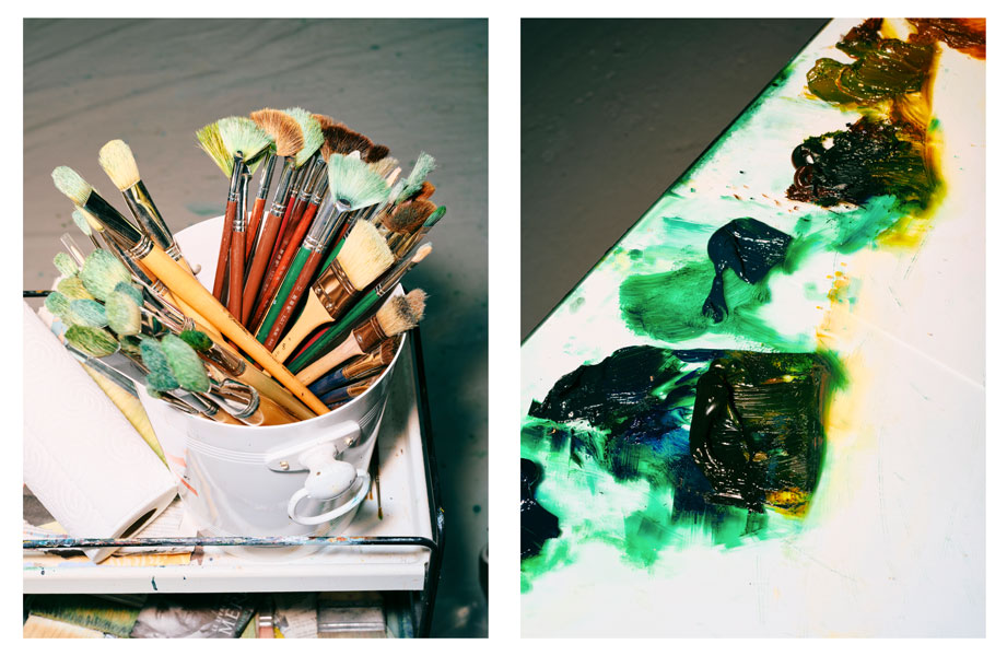 Gros plan sur l’atelier de l’artiste Xie Lei. À gauche, un seau rempli de pinceaux aux poils imprégnés de vert et de bleu. À droite, une palette couverte d’épaisses touches de peinture verte, jaune et noire, témoignant d’un travail pictural intense et expressif.